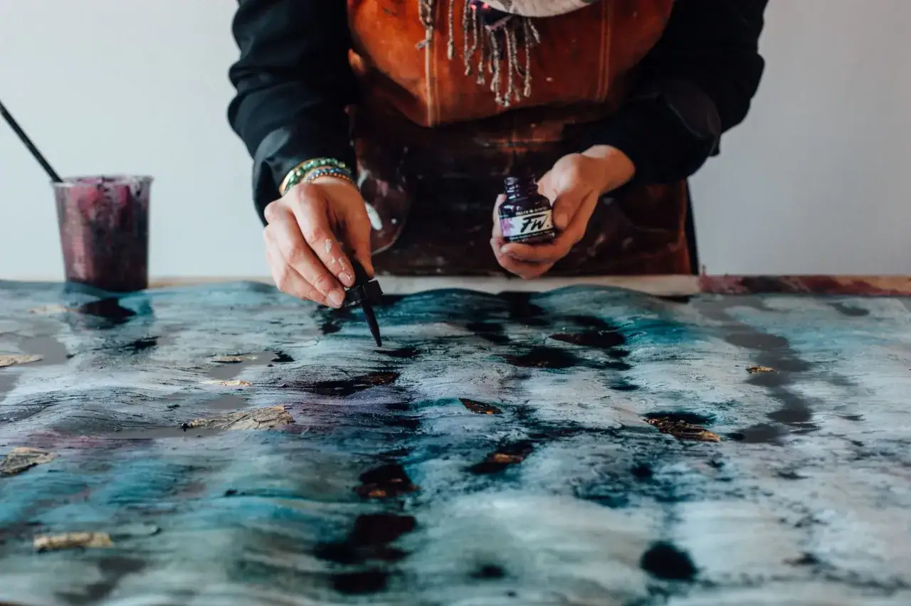 Manos de artista aplicando tinta p&uacute;rpura a una acuarela sobre lienzo con tonos azules y dorados, creando un efecto de olas.