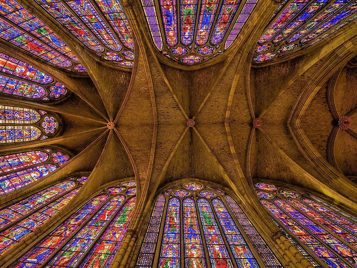 Techo abovedado de una catedral gótica, con vidrieras de colores vibrantes que iluminan el espacio. Las arte gótico características de arcos apuntados y bóvedas de crucería son evidentes.