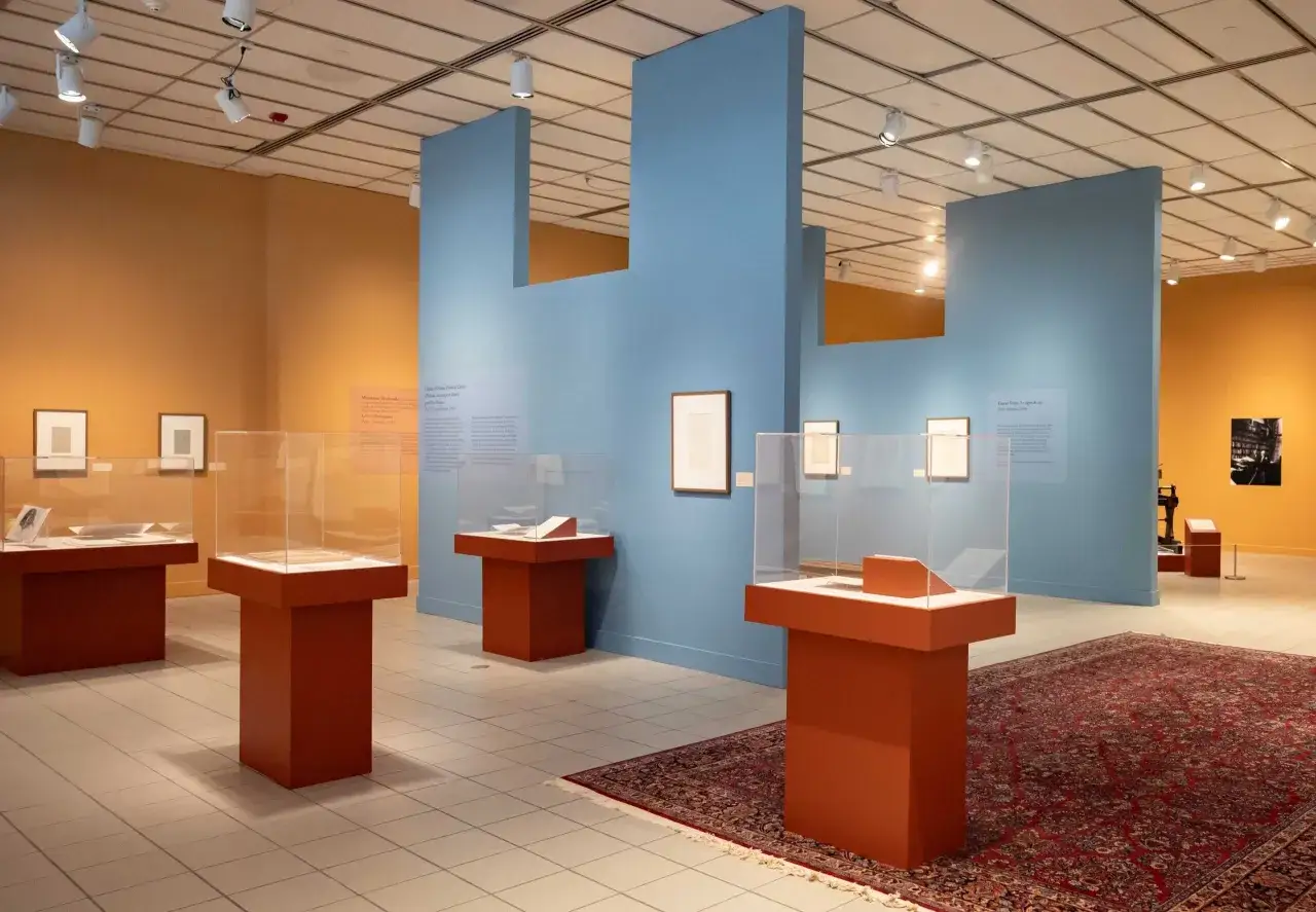 Exposici&oacute;n de obras de Henri Matisse, con vitrinas que exhiben dibujos y libros, y paredes de colores naranja y azul.