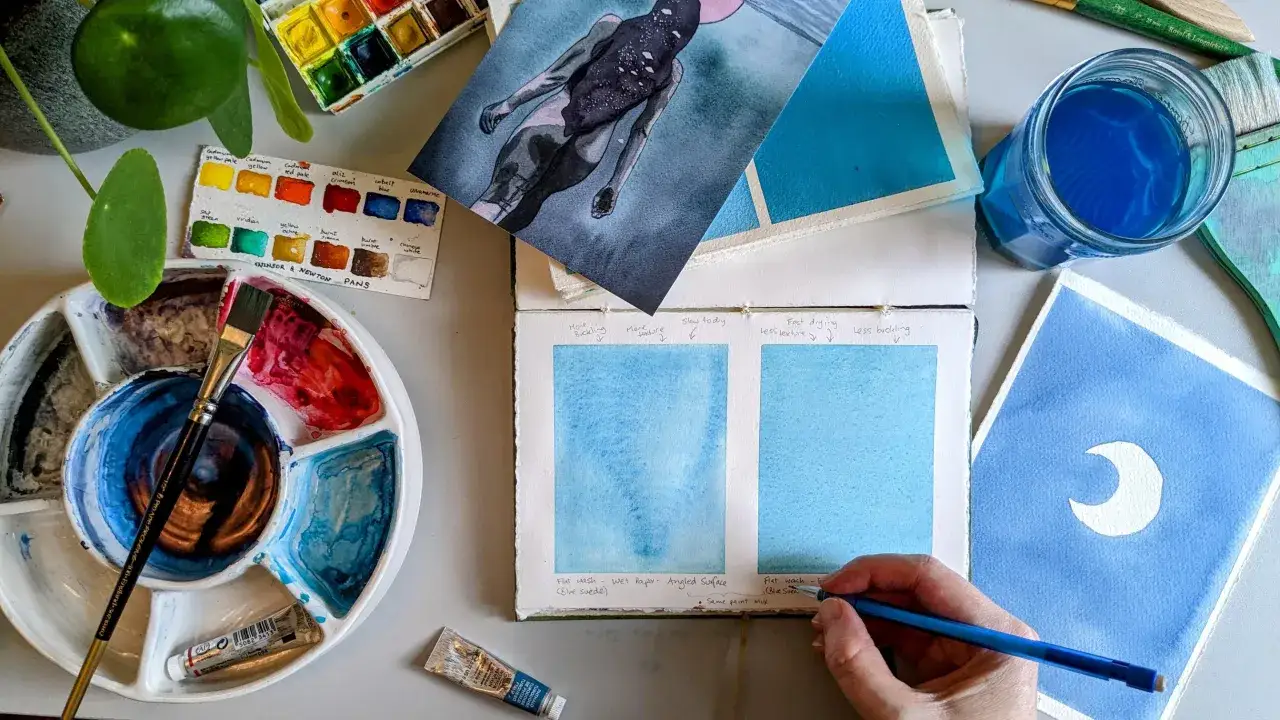Una mesa de trabajo con materiales para pintura en acuarela: paleta de colores, pinceles, tubos de pintura, un vaso con agua azul y varias obras en proceso.