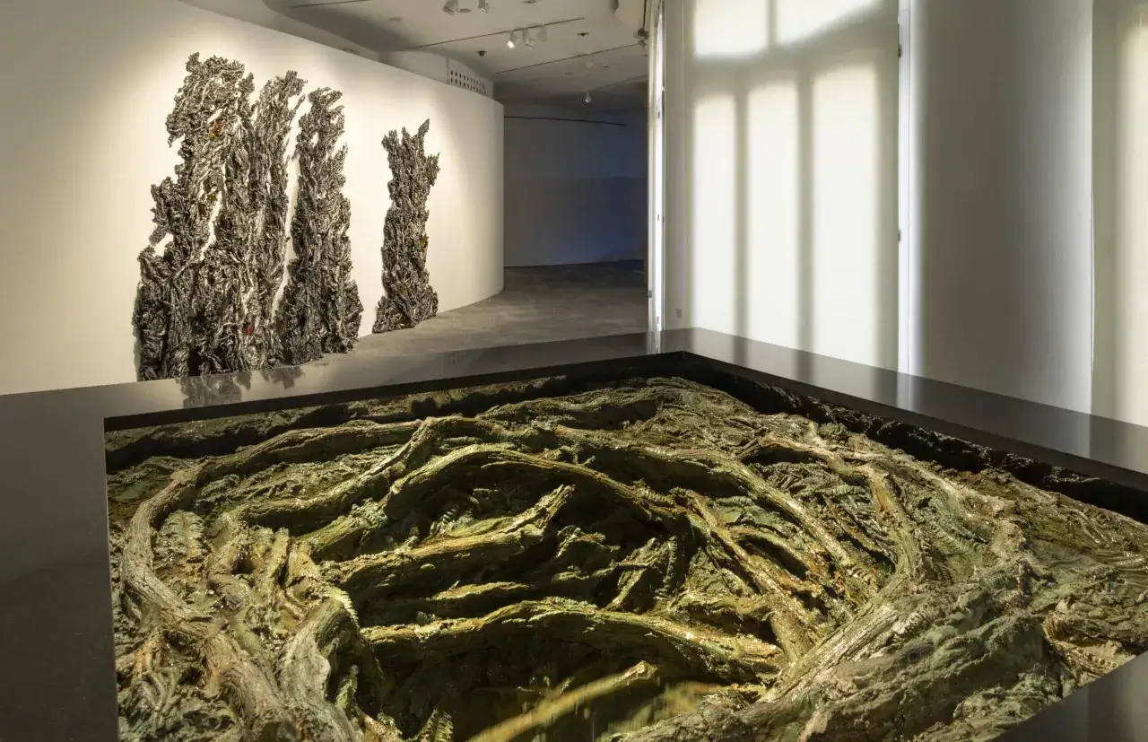 Instalaci&oacute;n de Cristina Iglesias: esculturas oscuras y texturizadas en la pared, y un intrincado paisaje de ra&iacute;ces verdes y marrones en un foso.