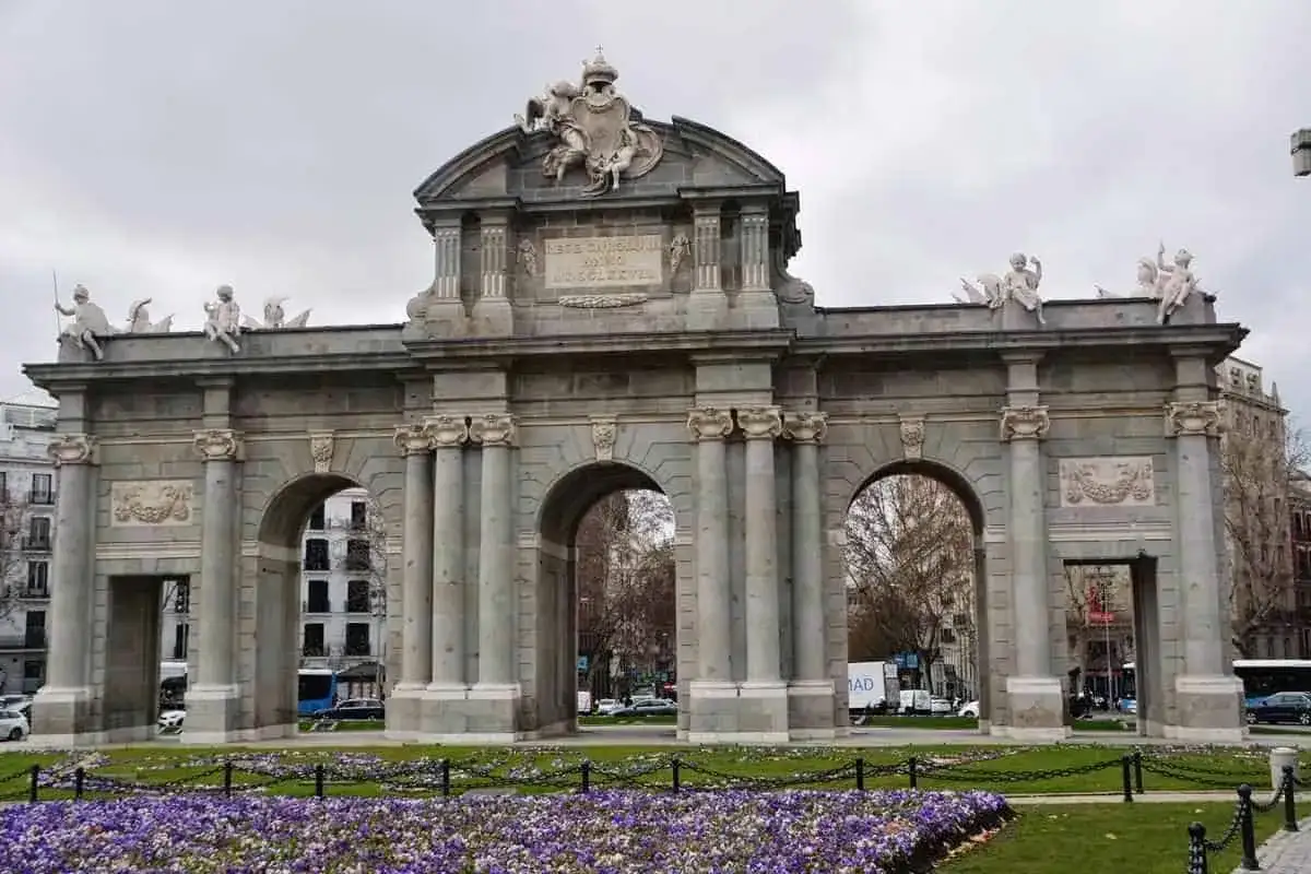 La Puerta de Alcal&aacute;, un monumento emblem&aacute;tico de Madrid, se alza majestuosa con sus arcos y esculturas, rodeada de un jard&iacute;n de flores moradas.