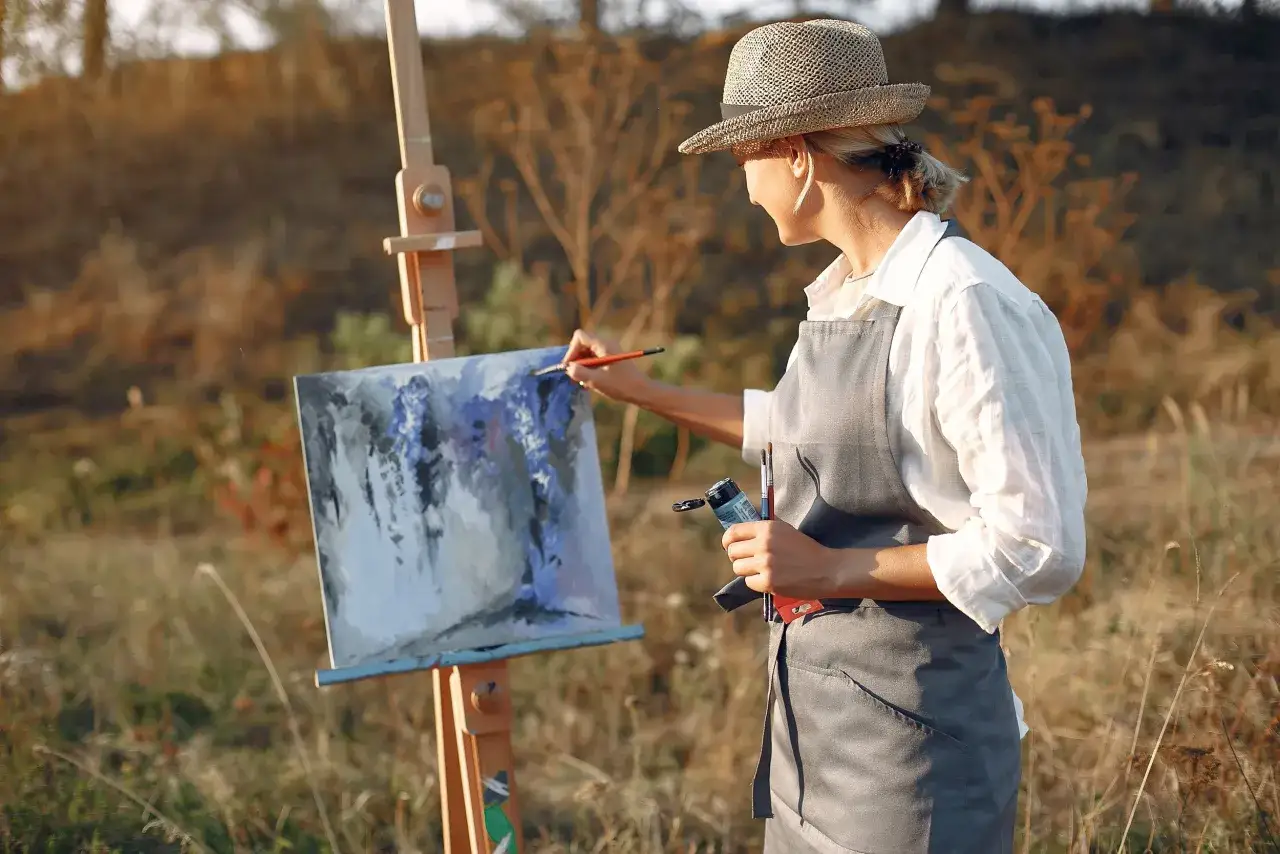 Mujer con sombrero pintando al aire libre, capturando la luz del atardecer en su lienzo.