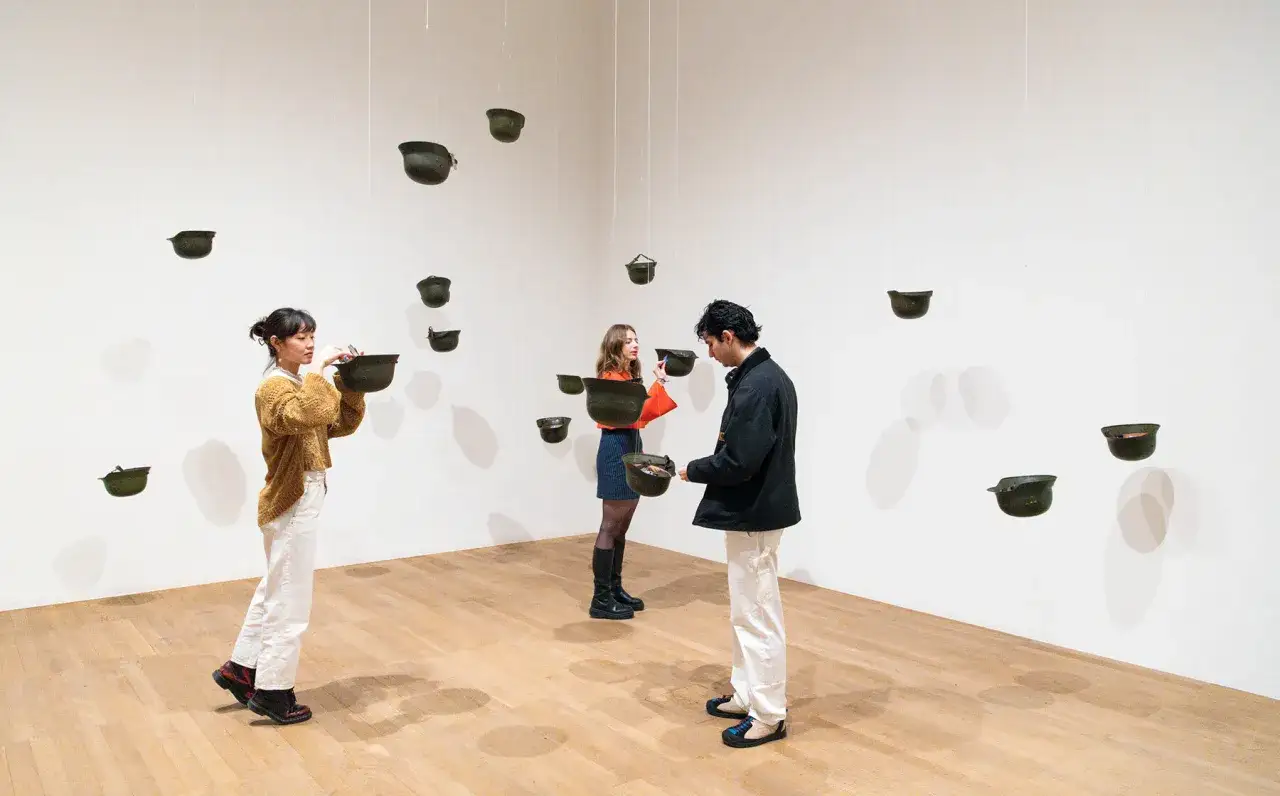 Personas interact&uacute;an con cascos militares suspendidos, parte de las obras de arte de Yoko Ono, en una galer&iacute;a.