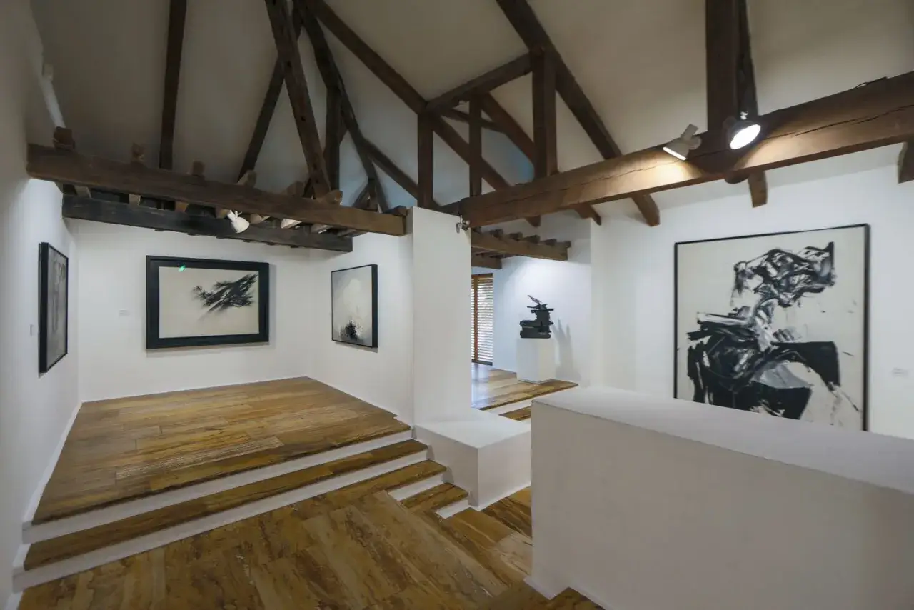 Galería de arte moderno con vigas de madera expuestas y suelo de madera. Destaca un gran cuadro de arte abstracto en blanco y negro.
